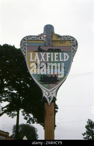 Suffolk. 1987 – Une photographie du signe du village de Laxfield dans le nord du Suffolk, en Angleterre. Le panneau est fait de bois sculpté et décoré de divers monuments et motifs locaux, y compris une locomotive à vapeur et des charrues. Laxfield est situé sur la route B1117 entre Ashfield Green et Ubbeston Green. Banque D'Images