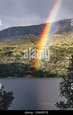 Un arc-en-ciel rayonnant forme un arc au-dessus d'un lac norvégien serein, se posant près de chalets traditionnels en rondins nichés parmi les collines d'Oppdal, en Norvège Banque D'Images