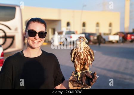 Jeune femme avec t-shirt noir et lunettes de soleil posant avec un oiseau de proie faucon arabe sur son gant en cuir de main, souriant Banque D'Images