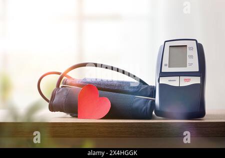 Moniteur de pression artérielle numérique bleu pour la maison sur table en bois avec découpe de coeur rouge pour la surveillance de la pression artérielle et fenêtre avec rideaux dans le BA Banque D'Images