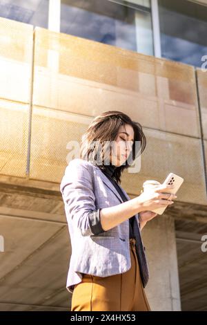 Jeune femme d'affaires marchant dans la rue tenant un téléphone portable et un verre de café, Une femme marchant dans la rue tenant un café en verre et utilisant Banque D'Images