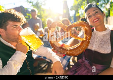 Homme buvant de la bière et une femme tenant un bretzel géant, tous deux dans le tracht allemand traditionnel, profitant d'une journée ensoleillée au jardin de bière ou à l'oktoberfest Banque D'Images