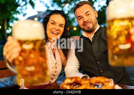 Heureux couple regardant et souriant à la caméra disent des acclamations ou des verres de clinking avec tasse de bière dans le jardin de bière bavarois ou oktoberfest Banque D'Images