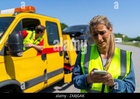 Femme en détresse avec un téléphone intelligent devant une dépanneuse et une voiture endommagée Banque D'Images