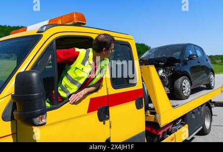 Opérateur de dépanneuse penché par la fenêtre du véhicule avec une voiture endommagée à l'arrière et vérifie si la voiture impliquée dans l'accident est correctement sécurisée Banque D'Images