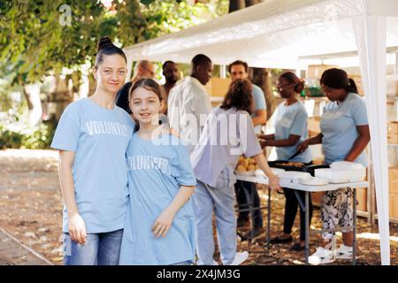 Portrait d'une mère et de sa fille, soutenant une initiative de lutte contre la faim dans une banque alimentaire en plein air. Deux femmes caucasiennes portant des t-shirts bleus écrits volontaires, prêts à aider les sans-abri. Banque D'Images