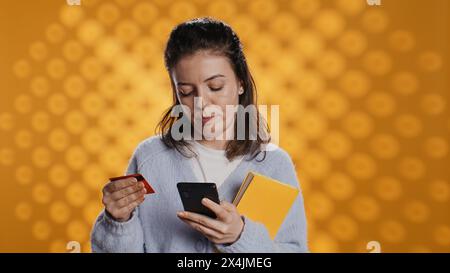 Femme tenant des livres, achetant plus avec le téléphone, véhiculant la joie du concept de lecture, fond de studio. Personne achetant des romans en ligne avec carte de crédit, montrant l'appréciation de la littérature, appareil photo B. Banque D'Images