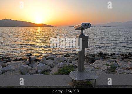 vue arrière gros plan de jumelles métalliques à pièces sur une jetée en béton sur la rive de la mer avec ciel au coucher du soleil en arrière-plan Banque D'Images