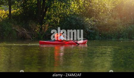 L'homme dans un kayak rouge pagaie sereinement sur une rivière calme, entouré d'un feuillage vert dense sous un soleil doré en été. Kayak Water Sports concept i Banque D'Images