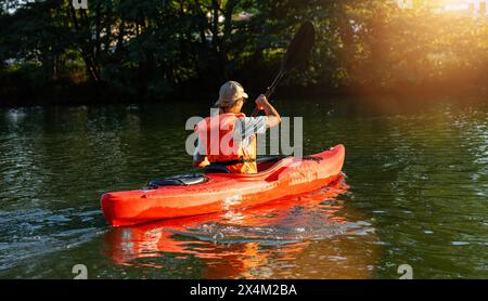 Femme en gilet de vie orange kayak sur une rivière tranquille avec une végétation luxuriante au coucher du soleil. Image concept de sports nautiques kayak Banque D'Images