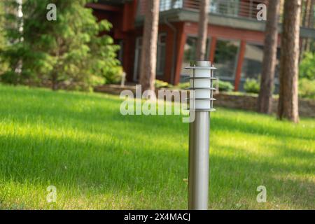 Un lampadaire haut et élégant en argent sur une pelouse verte vibrante près d'une maison en bois rouge. Le design moderne se marie à un environnement naturel. Arbres bien entretenus Banque D'Images