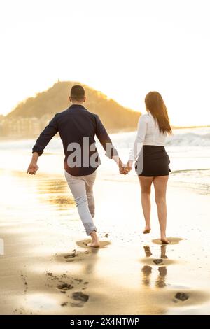 Photo verticale de la vue arrière d'un couple ludique courant se tenant la main sur le bord de la mer pendant le coucher du soleil Banque D'Images