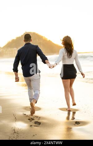 Photo verticale de la vue arrière d'un couple heureux tenant les mains courant le long d'une plage pendant le coucher du soleil Banque D'Images