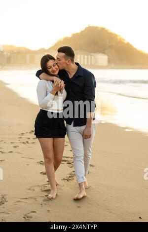 Photo verticale d'un jeune couple caucasien s'embrassant en marchant pieds nus le long d'une plage pendant le coucher du soleil Banque D'Images