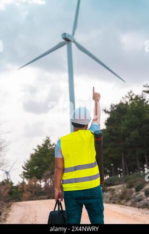 Photo verticale de la vue arrière d'un ingénieur adulte caucasien pointant vers une éolienne debout dans un parc d'énergie verte Banque D'Images