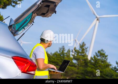 Technicien mûr masculin portant des vêtements réfléchissants et un casque rigide debout à côté de la voiture en utilisant un ordinateur portable travaillant dans un parc d'énergie éolienne avec des éoliennes Banque D'Images