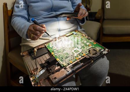 Réparations d'ordinateur, vieille femme dans les soixante-dix ans réparant son ordinateur portable, tenant la lumière du téléphone pour mieux voir, essayant de le réparer elle-même, amateur de bricolage Banque D'Images