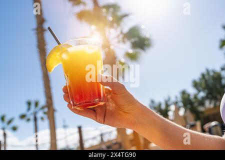 Gros plan d'une main tenant un cocktail tropical coloré sur fond de palmiers ensoleillés à l'hôtel caribbean Island Banque D'Images