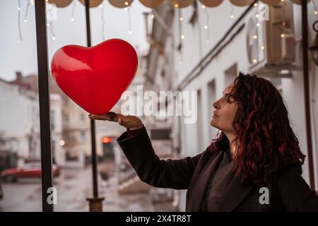Une femme tenant un ballon coeur rouge. La femme porte un manteau noir et a les cheveux roux. La scène se déroule dans une rue de la ville avec des voitures et un bâtiment dedans Banque D'Images