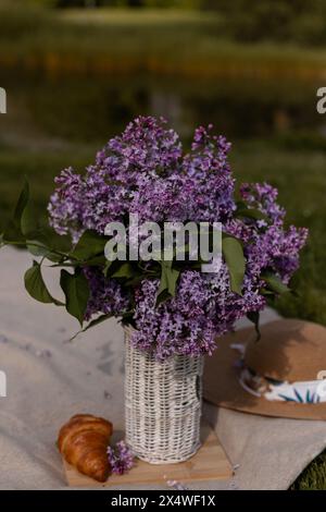 Bouquet de lilas dans un vase en osier, croissant et chapeau. Pique-nique de printemps. Photo de haute qualité Banque D'Images