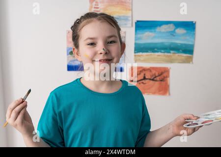 Portrait de petite adolescente mignonne avec pinceau et palette sur fond de ses dessins. Un jeune artiste au visage taché de peinture. Haute qualité Banque D'Images