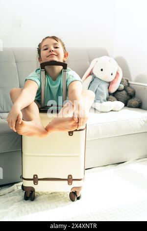 Petite fille - voyageuse, adolescente souriante - touriste assis avec des bagages, valise de voyage, faire le visage drôle, se préparer pour le voyage d'été et les vacances Banque D'Images