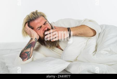 Homme barbu bâillant allongé sur un oreiller blanc doux. Bel homme dans un peignoir bâillant se réveiller dans un lit confortable à la maison le matin. Homme endormi au lit à Banque D'Images