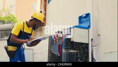 Électricien afro-américain effectuant des vérifications de fuites et d'autres réparations nécessaires pour éviter les pannes majeures. Technicien de maintenance habile vérifiant le climatiseur, écrivant les résultats sur le presse-papiers Banque D'Images