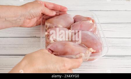 Cuisses de poulet désossées crues sans peau dans un récipient en plastique gros plan sur une planche de bois blanche dans les mains de la femme Banque D'Images