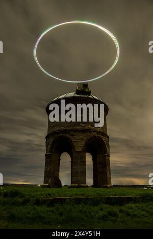 Vue nocturne énigmatique d'un ancien moulin à vent avec une traînée lumineuse de drone créant un effet de halo dans le ciel sombre Banque D'Images