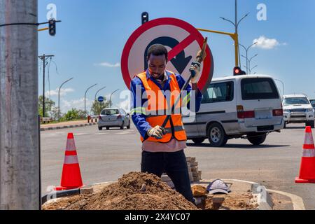 travailleur afro-américain avec une pioche creusant un fossé dans l'autoroute pour réparer le câblage sur une île à une intersection Banque D'Images