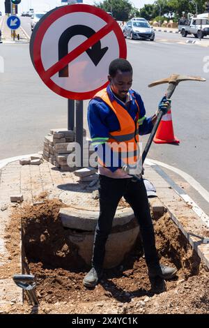 travailleur afro-américain avec une pioche creusant un fossé dans l'autoroute pour réparer le câblage sur une île à une intersection Banque D'Images