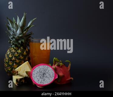 Un grand verre à facettes de jus de fruits sur un fond noir, à côté de morceaux et un ananas mûr avec pitaya. Gros plan. Banque D'Images