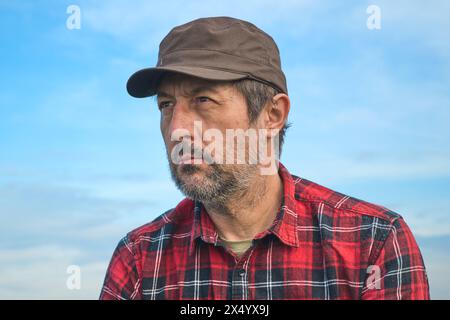 Portrait d'un fermier masculin sérieux confiant avec un chapeau brun et une chemise posée rouge contre un ciel bleu, mise au point sélective Banque D'Images