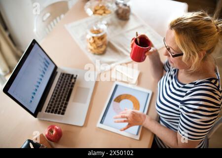 Femme plus âgée travaillant à domicile, tapant sur ordinateur portable, regardant la statistique dans la tablette. Le retraité gagne de l'argent supplémentaire pendant sa retraite. Banque D'Images