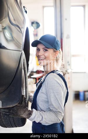 Mécanicien automobile féminin réparant, entretenant la voiture. Belle femme debout dans un garage, portant des combinaisons bleues. Banque D'Images