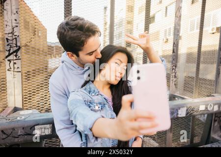 Couple assis sur un pont sur un chemin de fer urbain souriant et prenant un selfie avec un téléphone. Couple interracial traînant et prenant un selfie. Banque D'Images