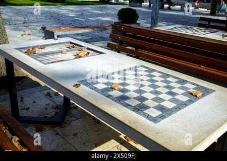 Les tables d'échecs attendent les joueurs dans un parc de la ville. Sérénité automnale. La lumière du soleil baigne les tables d'échecs vides et les bancs entourés de feuilles tombées sur un au clair Banque D'Images