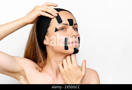 Rajeunissement facial et thérapie alternative anti-âge. Modèle de beauté femme Brunette appliquant des bandes faciales kinesio sur son visage devant le dos blanc Banque D'Images