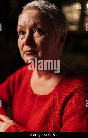 Gros plan sur la tête d'une femme âgée aux cheveux gris attrayante dans un pull rouge décontracté assis à table dans une pièce sombre par la lumière de la bougie allumée Banque D'Images
