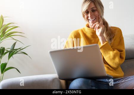 Femme heureuse en pull jaune à l'aide d'un ordinateur portable pour les médias sociaux sur le canapé, plante d'intérieur dans backgroung Banque D'Images