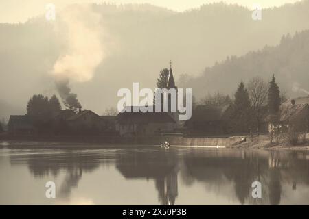 Scène matinale froide sur une rive du lac avec de la fumée venant d'une cheminée dans une maison de village. Banque D'Images