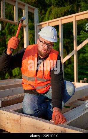 Charpentier construisant une maison à ossature en bois de deux étages près de la forêt. Homme barbu portant des lunettes martelant des clous dans la structure. Le projet incarne le concept de construction écologique moderne. Banque D'Images