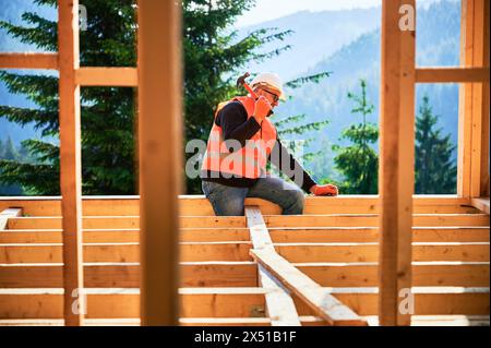 Carpenter construisant une maison en bois de deux étages près de la forêt. Homme martelant des clous dans la structure, portant un casque de protection et un gilet de construction. Concept de construction écologique moderne. Banque D'Images