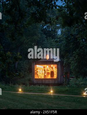 Le pavillon Apiaire vu au crépuscule de l'autre côté d'un champ, entouré d'arbres. Les lumières intérieures sont dorées montrant l'arrière du bâtiment, les fosses de sièges et l'exhib Banque D'Images