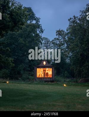 Le pavillon Apiaire vu au crépuscule de l'autre côté d'un champ, entouré d'arbres. Les lumières intérieures sont dorées montrant l'arrière du bâtiment, les fosses de sièges et l'exhib Banque D'Images