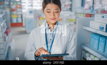 Pharmacie Drugstore : Portrait de belle pharmacienne asiatique utilise Digital Tablet Computer et sourit charmablement, derrière ses étagères pleines de paquets de médecine. Gros plan moyen Banque D'Images