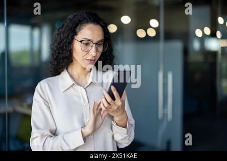 Une femme d'affaires hispanique concentrée en lunettes utilise son smartphone, lisant éventuellement un message important ou vérifiant les mises à jour de l'entreprise dans un environnement de bureau moderne. Banque D'Images