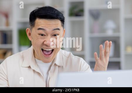 Un homme adulte asiatique enthousiaste exprimant la joie et le succès tout en travaillant à la maison. Il est visiblement joyeux, faisant des gestes avec ses mains, dans une pièce très éclairée. Banque D'Images