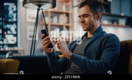 Homme caucasien excité à l'aide d'un smartphone tout en se reposant sur un canapé dans le salon élégant. Homme heureux souriant à la maison et discutant avec des collègues et des clients sur Internet. Utilisation des réseaux sociaux. Banque D'Images